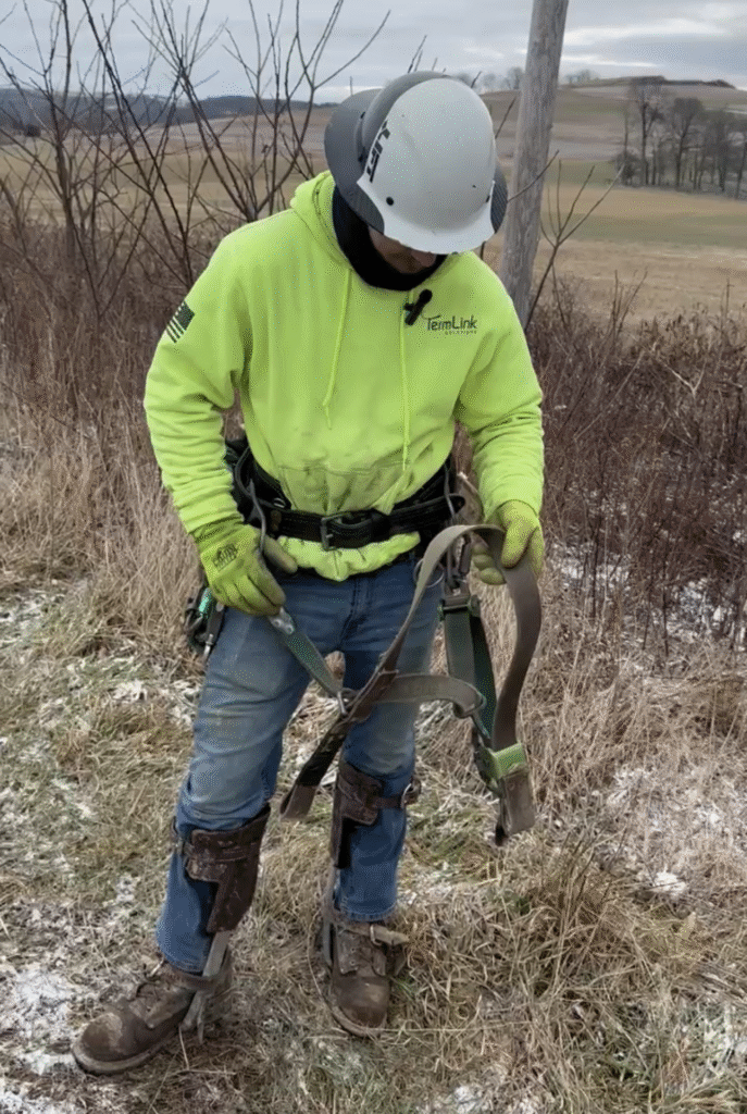 Fiber worker inspecting safety harness before climbing utility pole.